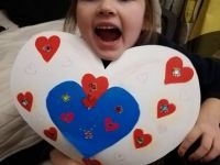 Czwarte zdjęcie przedstawia Laurę Kozak, dziewczynkę z rozweseloną buzią która trzyma białe serce na którym naklejone są mniejsze czerwone serca z cekinami. Całość serca uzupełnia mniejsze niebieskie serduszko na środku.